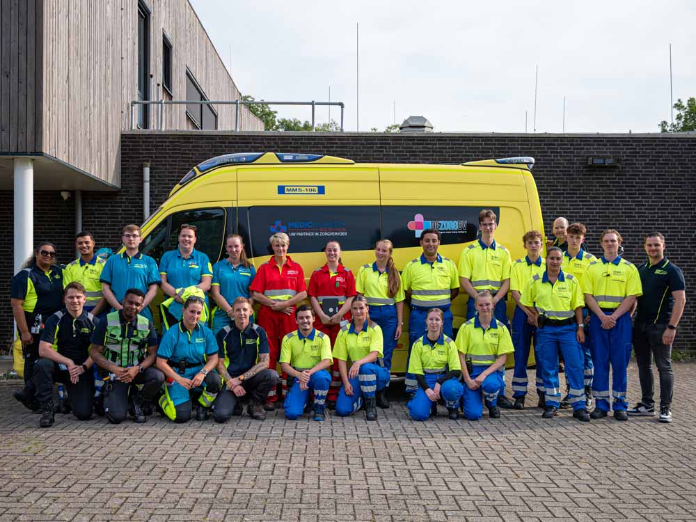 Groepsfoto van EHBO- en evenementenzorgmedewerkers voor een ambulancevoertuig.