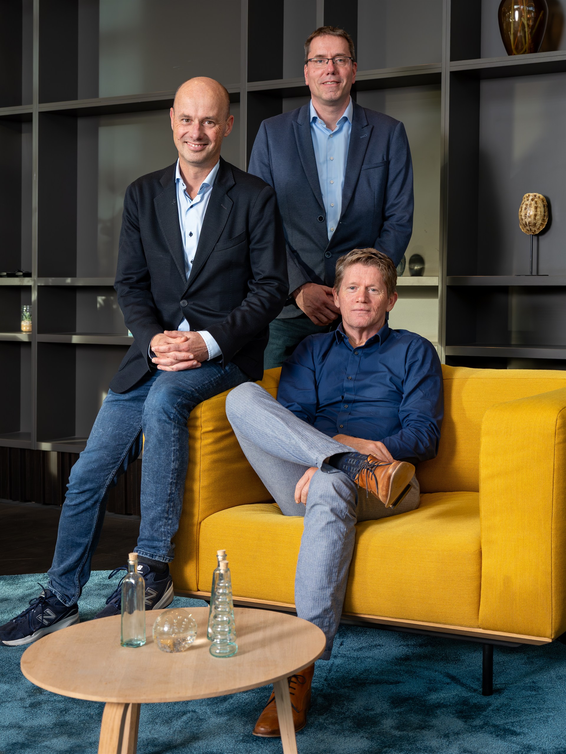 Zakelijke groepsfoto van drie mannen in een modern kantoor bij een gele bank.