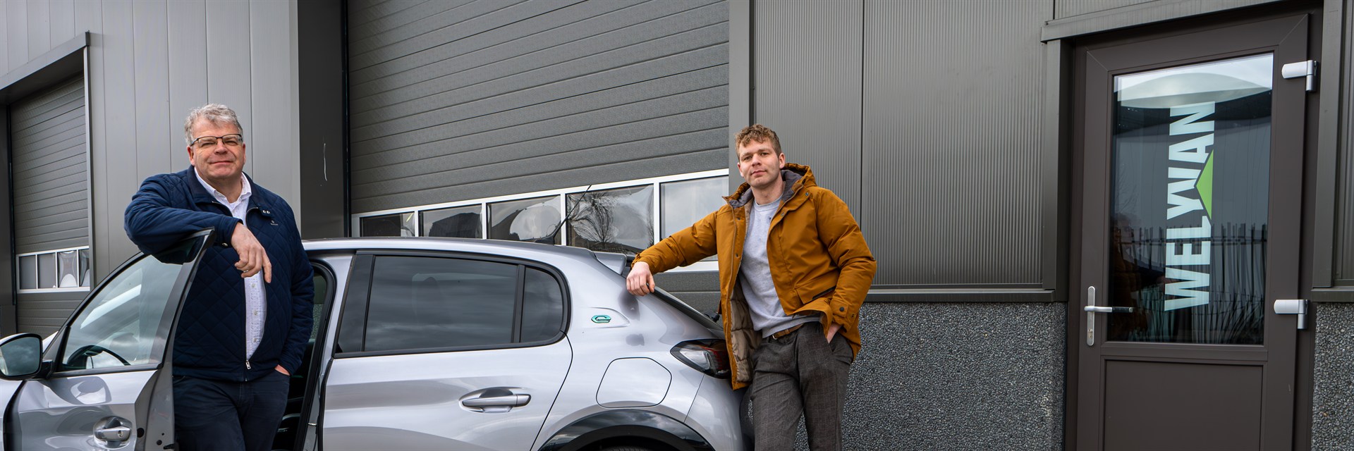Twee medewerkers van Prefab Care bij een auto voor de werkplaats van het bedrijf.